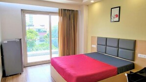 A hotel room with a bed, a small refrigerator, a sliding glass door to the balcony, and a framed picture on the wall. - Kommune Living (Gurugram)