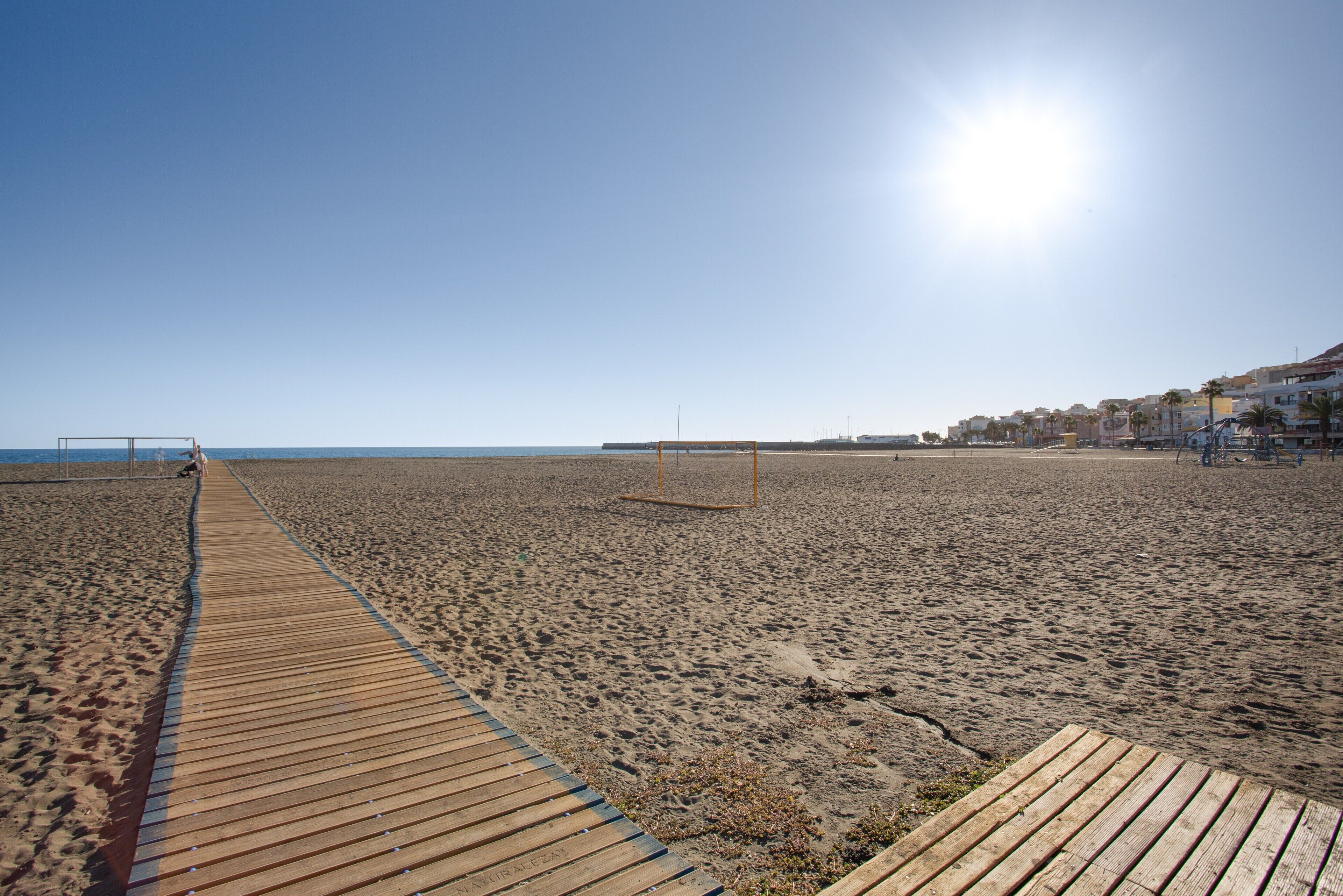 Una playa con un paseo marítimo de madera que llega al agua, cielo despejado y edificios al fondo.