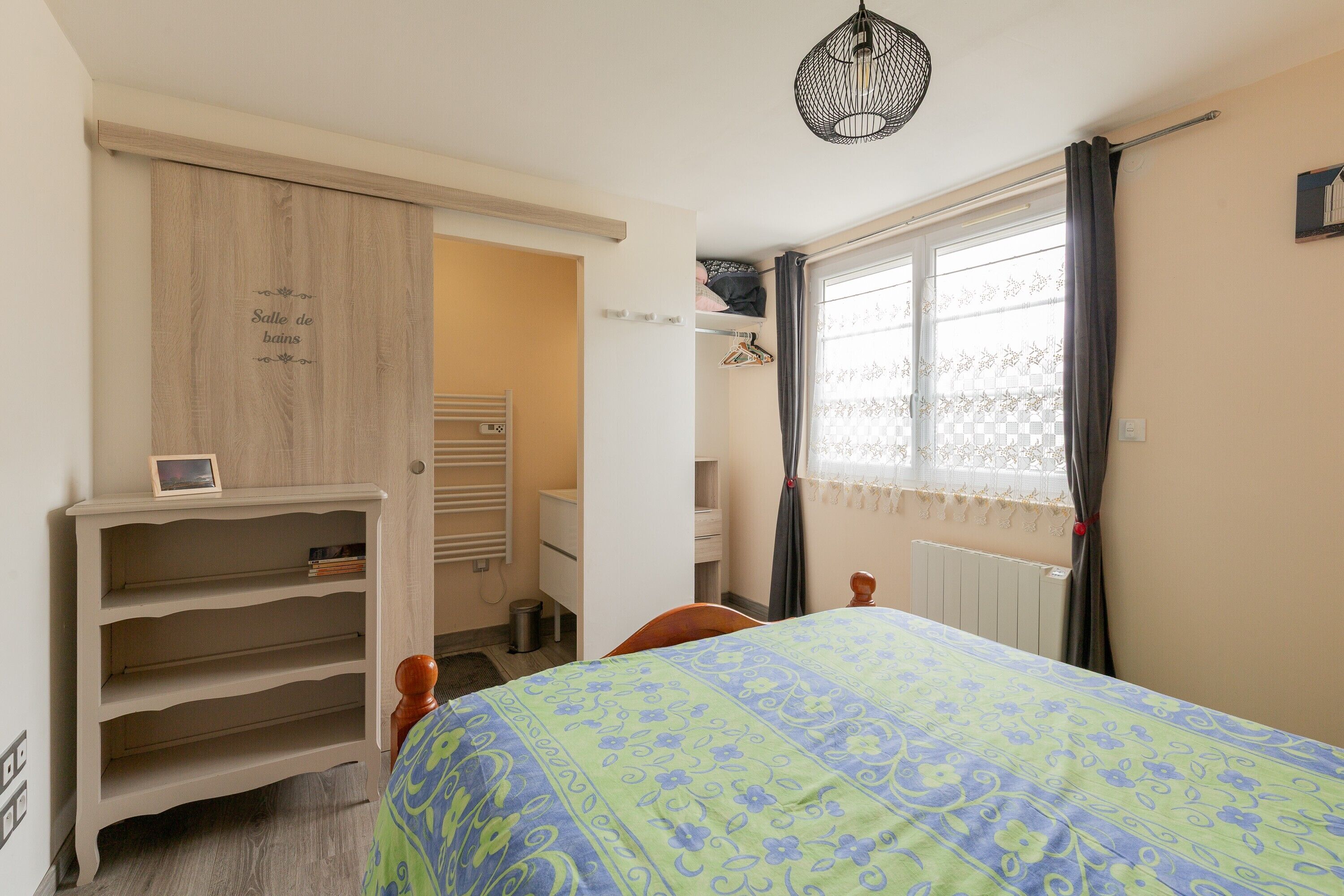 Um quarto com uma cama, um guarda-roupa, um radiador, uma janela com cortinas e um candeeiro suspenso.