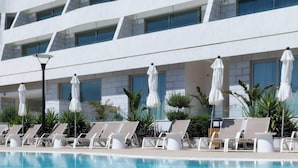 A poolside area with lounge chairs and umbrellas, a modern building with large windows, and a clear blue pool. - By Edom - דירה נוף מטריף לים במתחם סי סייד (Eilat)
