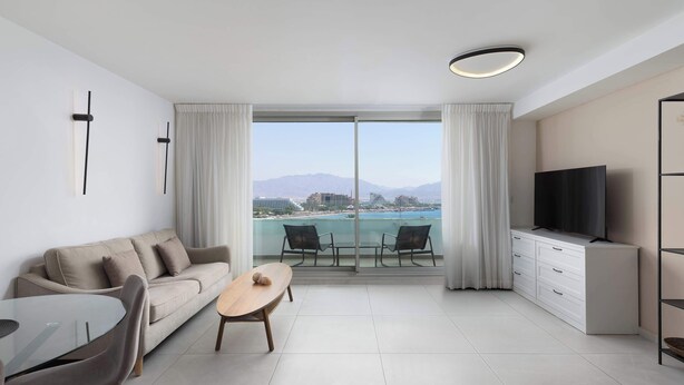 A modern living room with a sofa, dining table, chairs, a TV, and a balcony with a city view. - By Edom - דירה נוף מטריף לים במתחם סי סייד (Eilat)