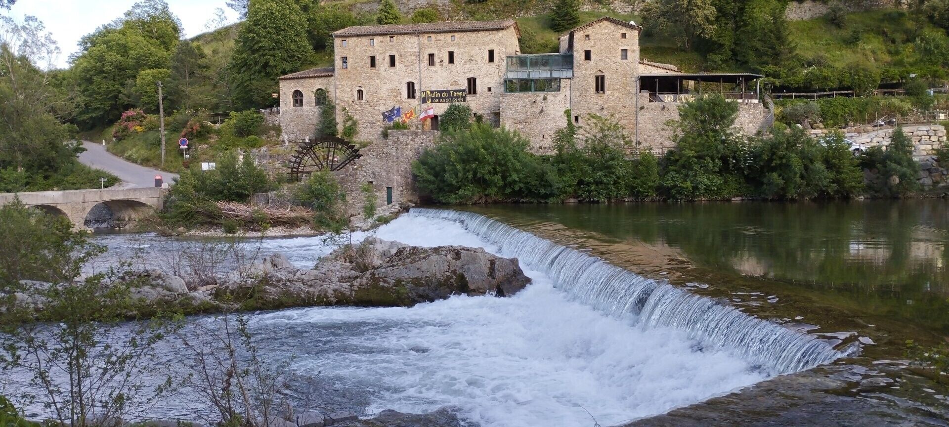 Un edificio histórico de piedra con un elemento acuático en primer plano, un puente y abundante vegetación.