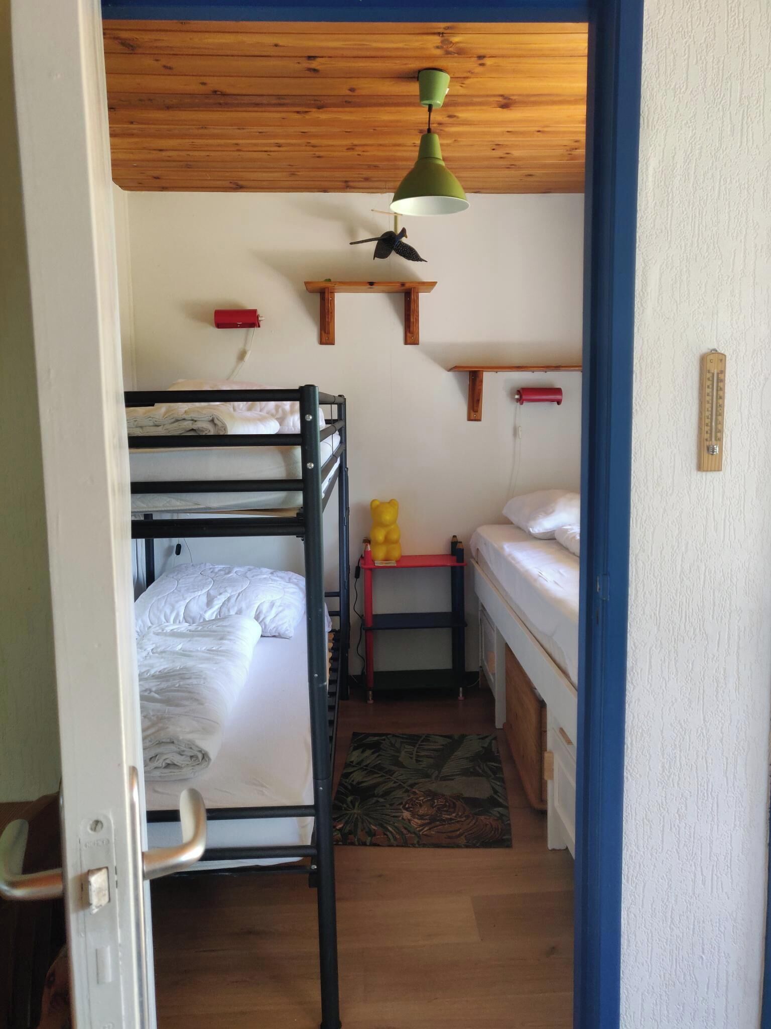 Un dormitorio con literas, una mesita con un osito de peluche, un calefactor rojo y un termómetro en la pared.