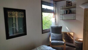 Ein Zimmer mit einem Bett, einem Sessel, einem Fenster mit Blick auf Grünflächen, einem Bücherregal und einem gerahmten Bild an der Wand.