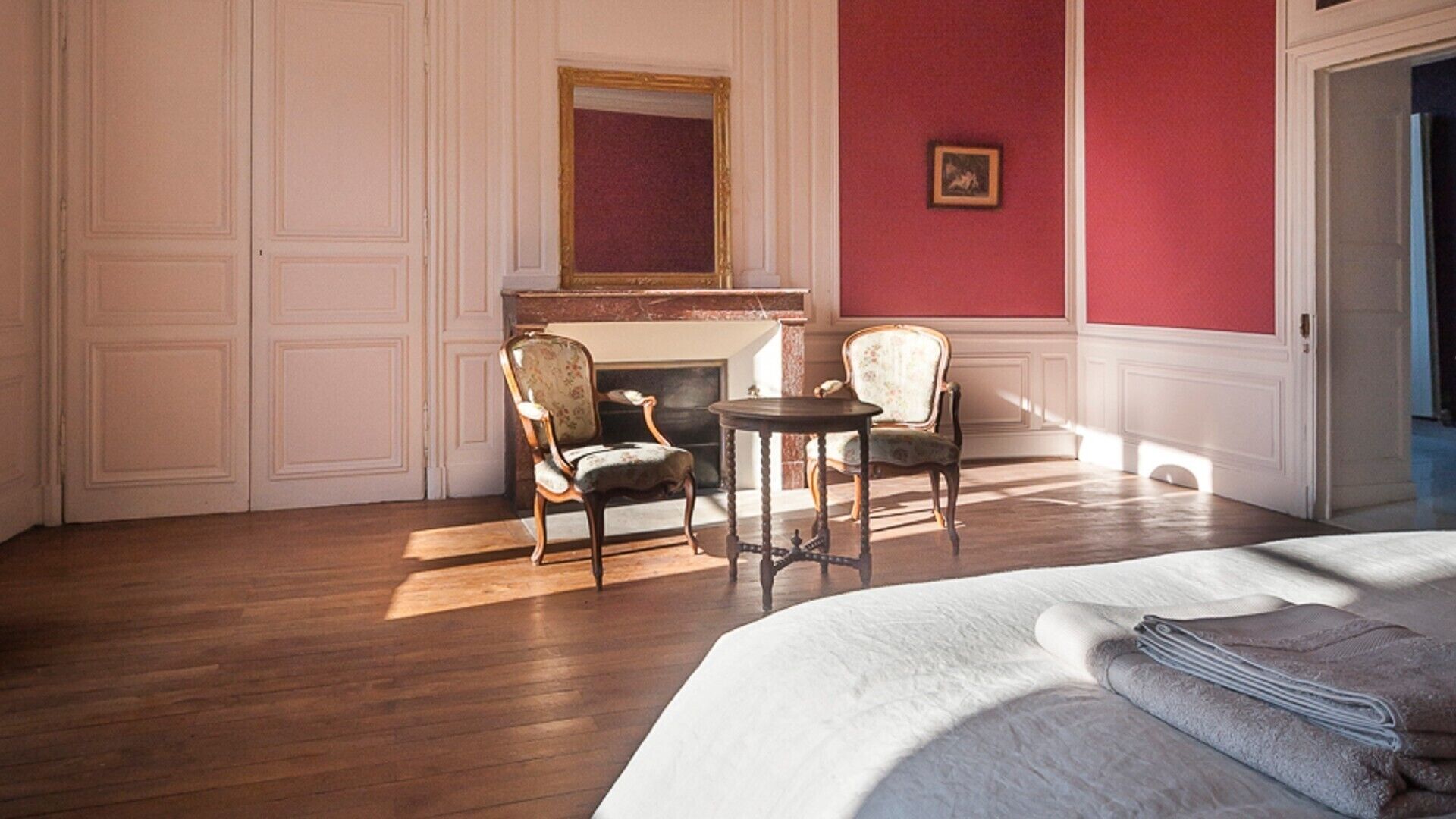 A room with a bed, two chairs, a small table, a fireplace, and a mirror on the wall.