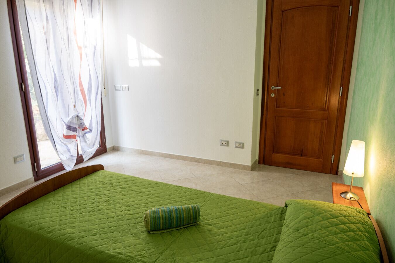 Een slaapkamer met een groen gewatteerde sprei, een houten deur, een nachtkastje met een lamp en een raam met dunne gordijnen.