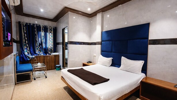 Une chambre d’hôtel moderne dotée d’un grand lit, d’une table de chevet, d’une commode, d’une salle de bain avec douche et d’un coin salon avec un banc et des rideaux.