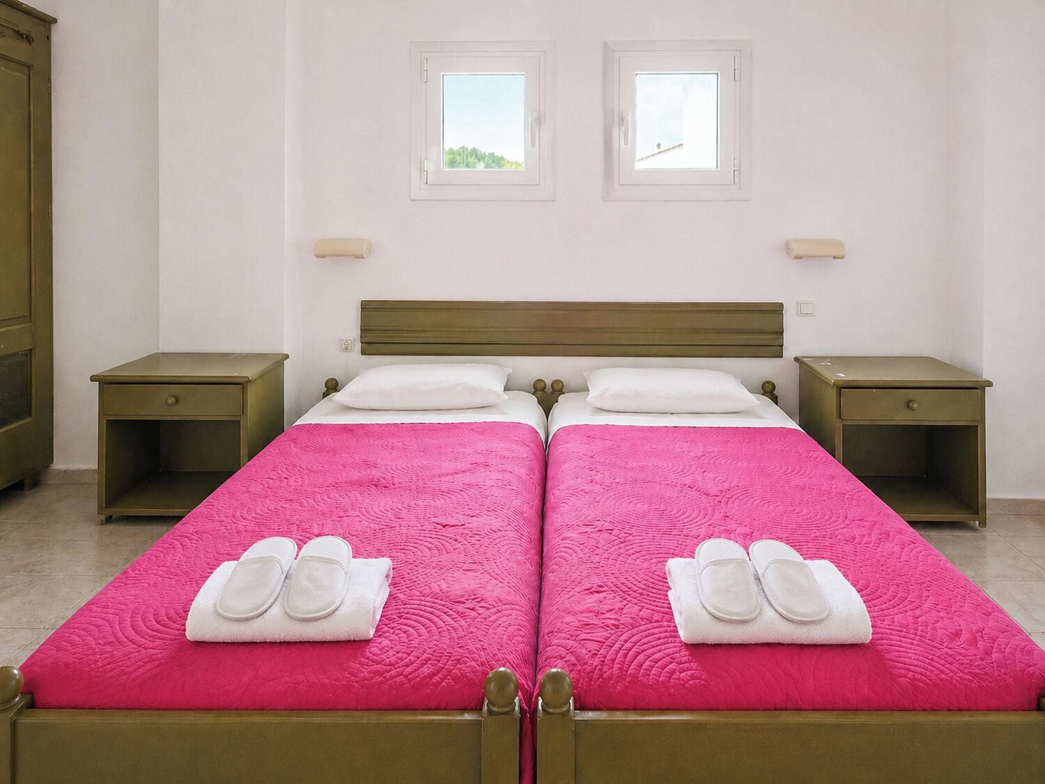 Une chambre double avec deux lits, chacun recouvert d’une couette rose et agrémenté de serviettes blanches. La chambre est équipée de deux tables de chevet et de deux fenêtres.