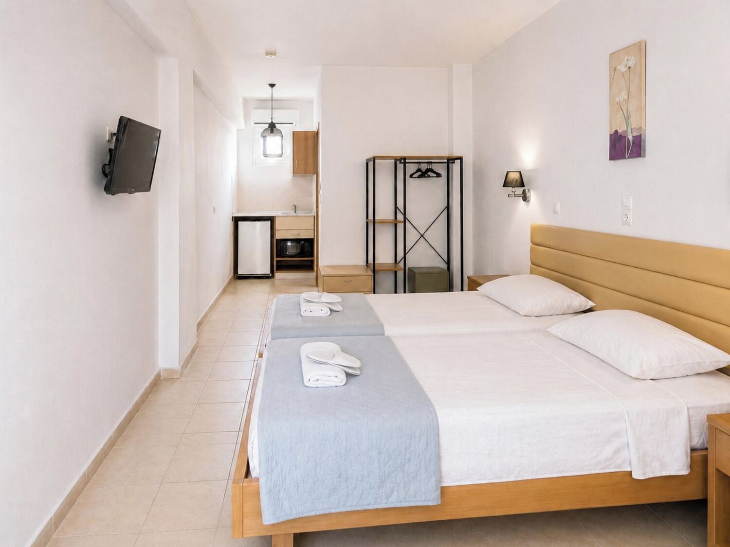A hotel room with a bed, bedside tables, a TV mounted on the wall, a small kitchenette, and a bathroom visible in the background.