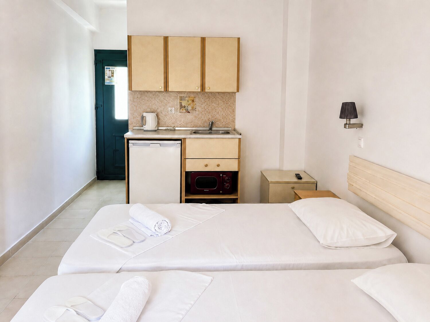 Una habitación de hotel con una cama, una kitchenette pequeña, una mesita de noche y una lámpara.