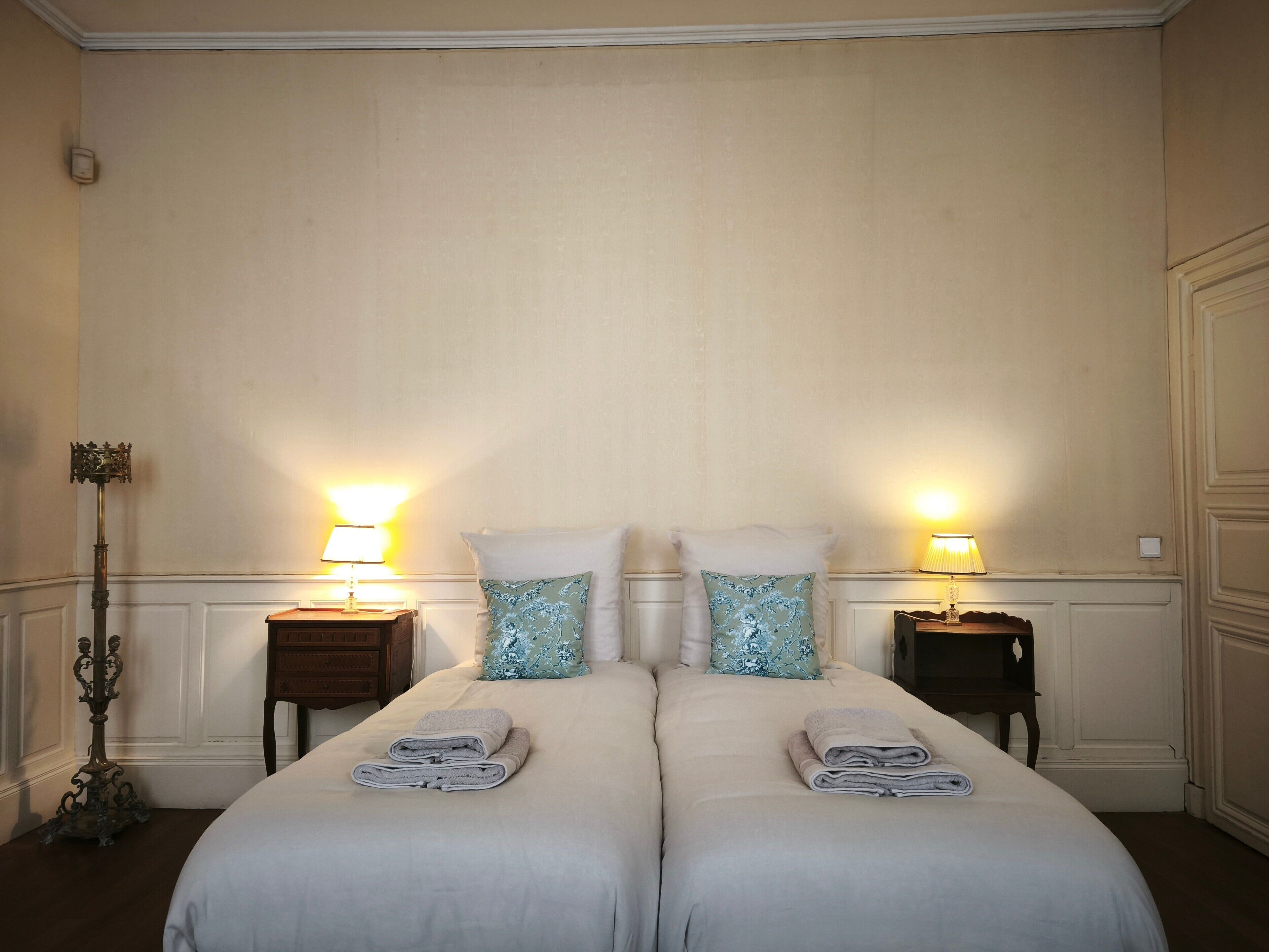 A hotel room with two beds, each with a pillow and a folded towel. There are bedside lamps and a floor lamp.