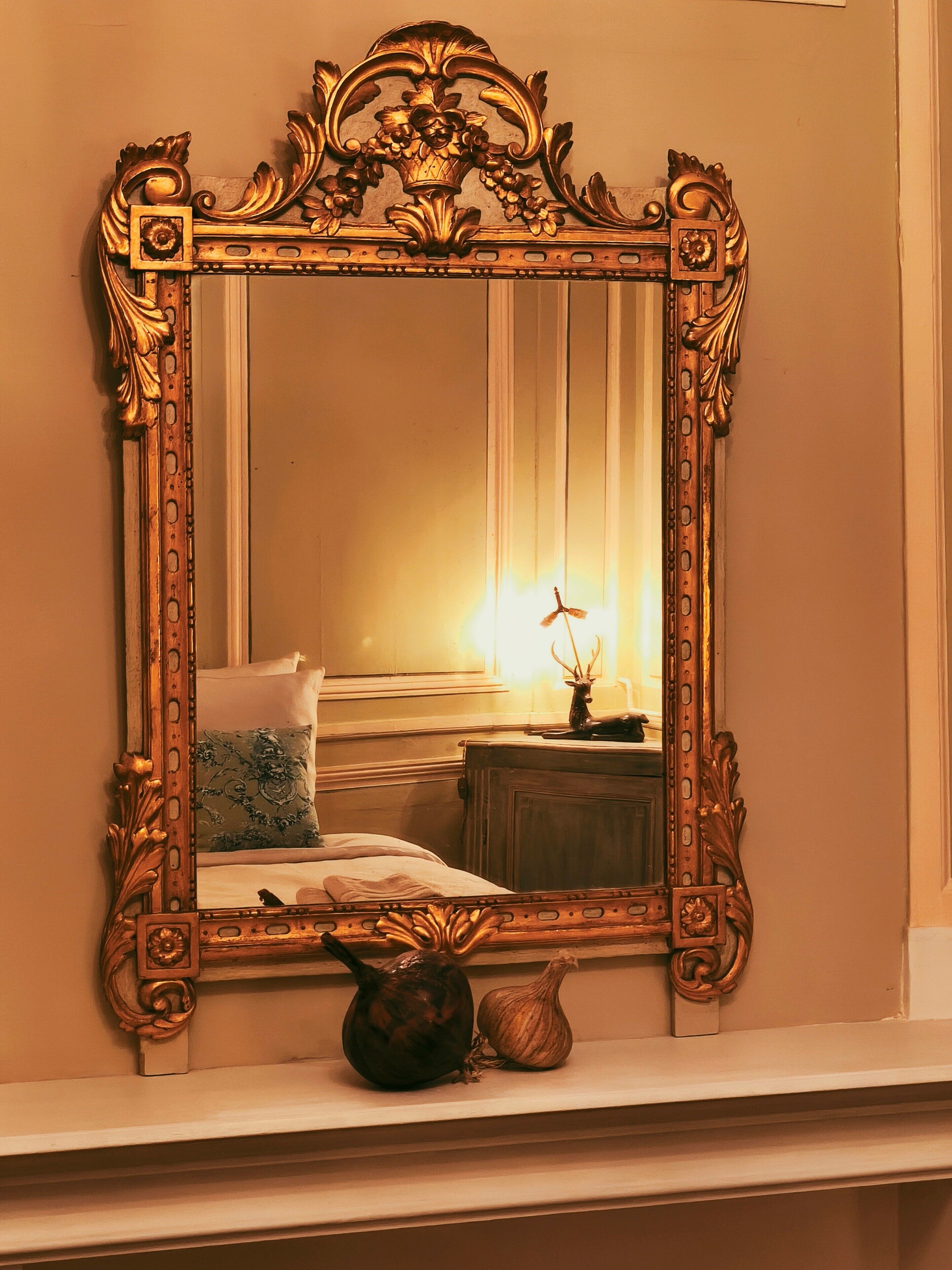 A mirror with ornate gold detailing, reflecting a bed with a pillow and a nightstand with a lamp and a vase.