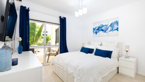 A bedroom with a large bed, white nightstands, a desk with a vase, and a view of greenery outside. - Lounge Garden Apartment (Loulé)