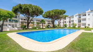 A swimming pool surrounded by a well-maintained lawn and trees, with a multi-story building in the background. - Lounge Garden Apartment (Loulé)