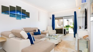 A modern living room with a beige sofa, a glass coffee table, and a flat-screen TV. There is a dining area with a white table and chairs, and a view of the outdoors through a glass door. - Lounge Garden Apartment (Loulé)