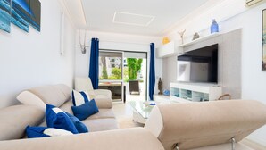 A modern living room with a beige sofa, a glass coffee table, and a flat-screen TV on a white entertainment unit. - Lounge Garden Apartment (Loulé)
