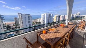 A balcony with a wooden table, chairs, and a view of the beach and cityscape. - Ocean View Quarteira (Loulé)