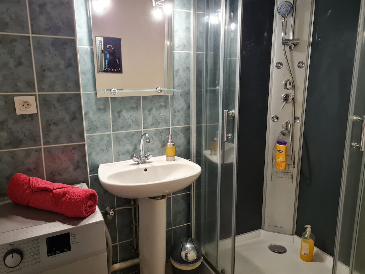 Une salle de bain avec un lavabo blanc, une serviette rouge, une cabine de douche en verre et un miroir au-dessus du lavabo.