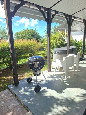 Een overdekt terras met een barbecue, tuinmeubilair en uitzicht op een tuin.