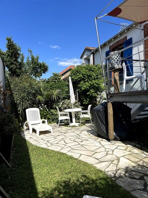 Une terrasse avec des meubles d’extérieur, une table abritée et un bâtiment aux volets bleus.