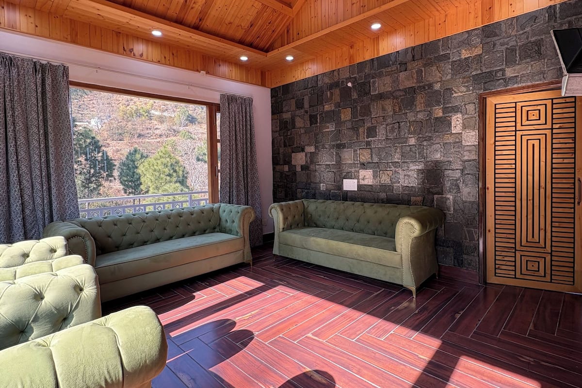 A room with a wooden ceiling, stone wall, and a large window with curtains. There are two green sofas and a wooden cabinet.