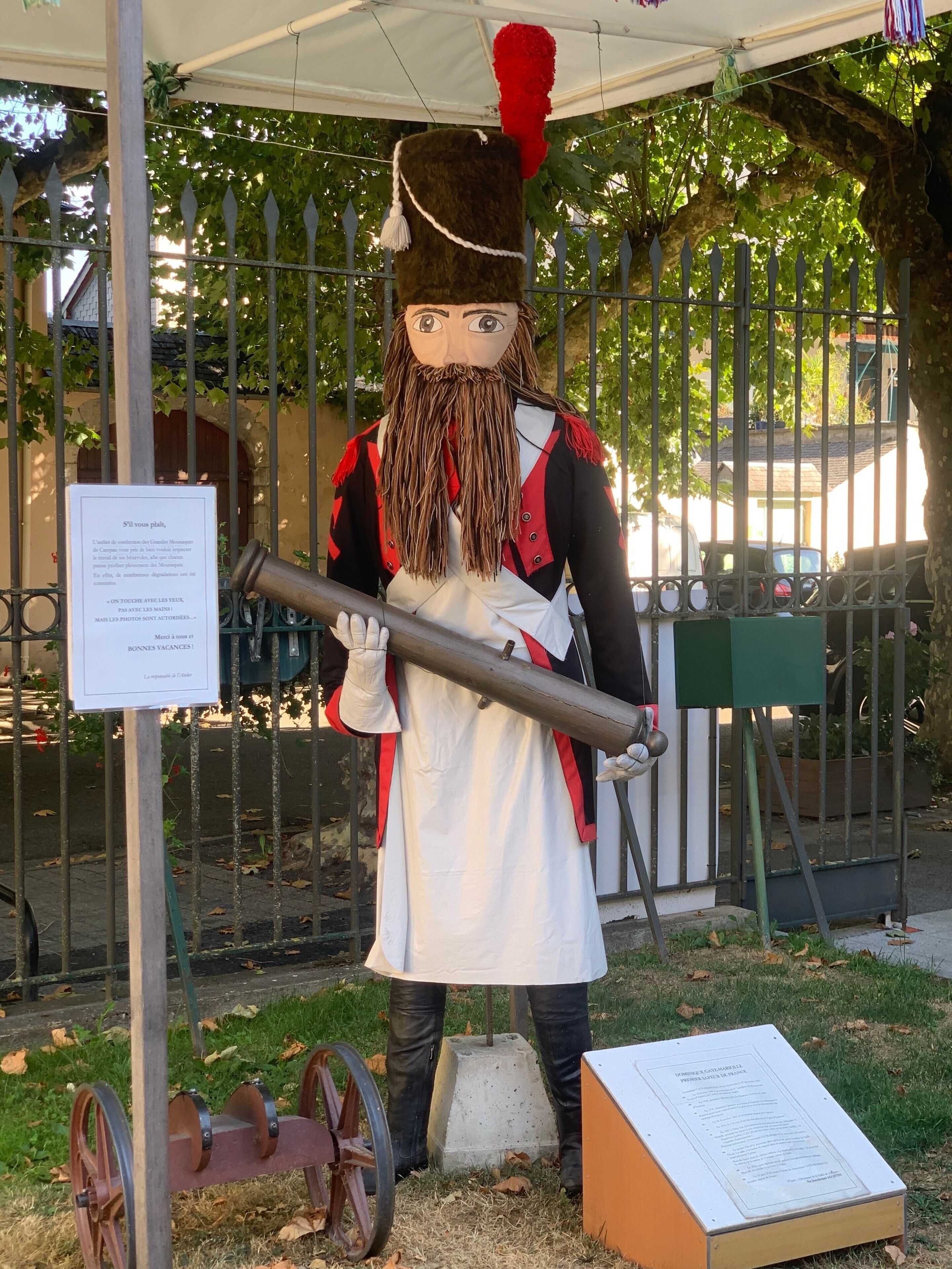 Una estàtua d'un personatge que sosté un objecte gran, situada al costat d'un senyal amb text, sota una marquesina amb una tanca al fons.