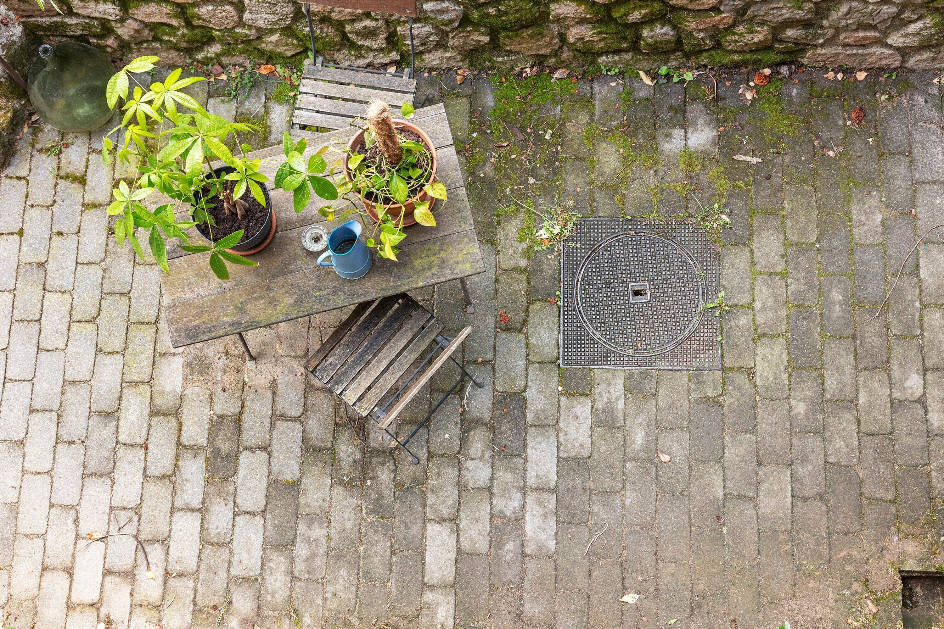Een houten tafel met potplanten, een blauwe mok en een metalen rooster op een terras met bakstenen bestrating.