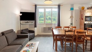 A modern living room with a sofa, a coffee table with magazines, a dining table with chairs, a TV mounted on the wall, and a kitchen area with cabinets and a microwave. - “Grand Gîte” with Mountain View, Shared Pool, and Wi-Fi (Saint-Jean du Gard)