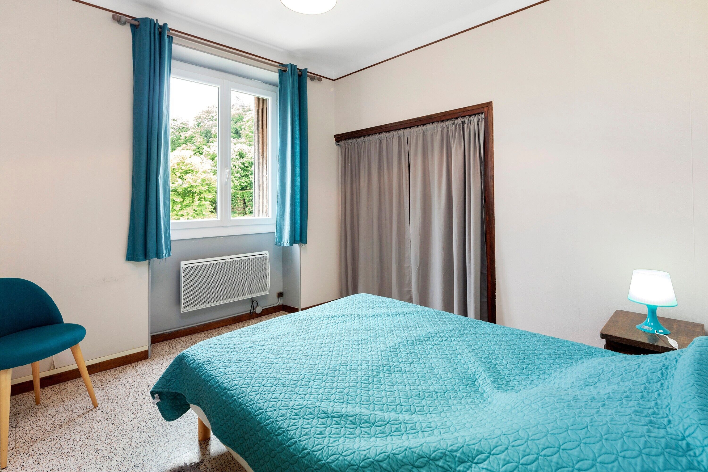 Une chambre à coucher comprenant un lit, une chaise, une fenêtre avec des rideaux, un radiateur et une table de chevet avec une lampe.
