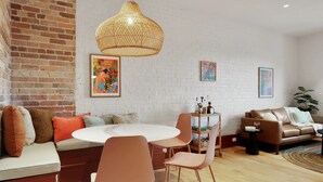 Una sala moderna con pared de ladrillo, mesa redonda para comer, sillas naranjas, un sofá de cuero marrón y una lámpara colgante tejida.