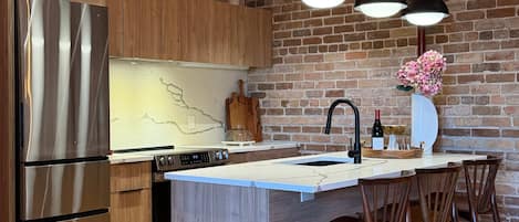Una cocina moderna con un refrigerador de acero inoxidable, armarios de madera, una encimera de mármol y una pared de ladrillo. Hay tres lámparas colgantes, un lavamanos y taburetes de bar.