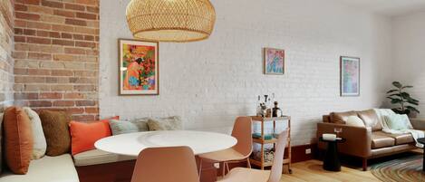 A modern living room with a brick wall, white brick wall, round dining table, orange chairs, brown sofa, and a hanging light fixture.