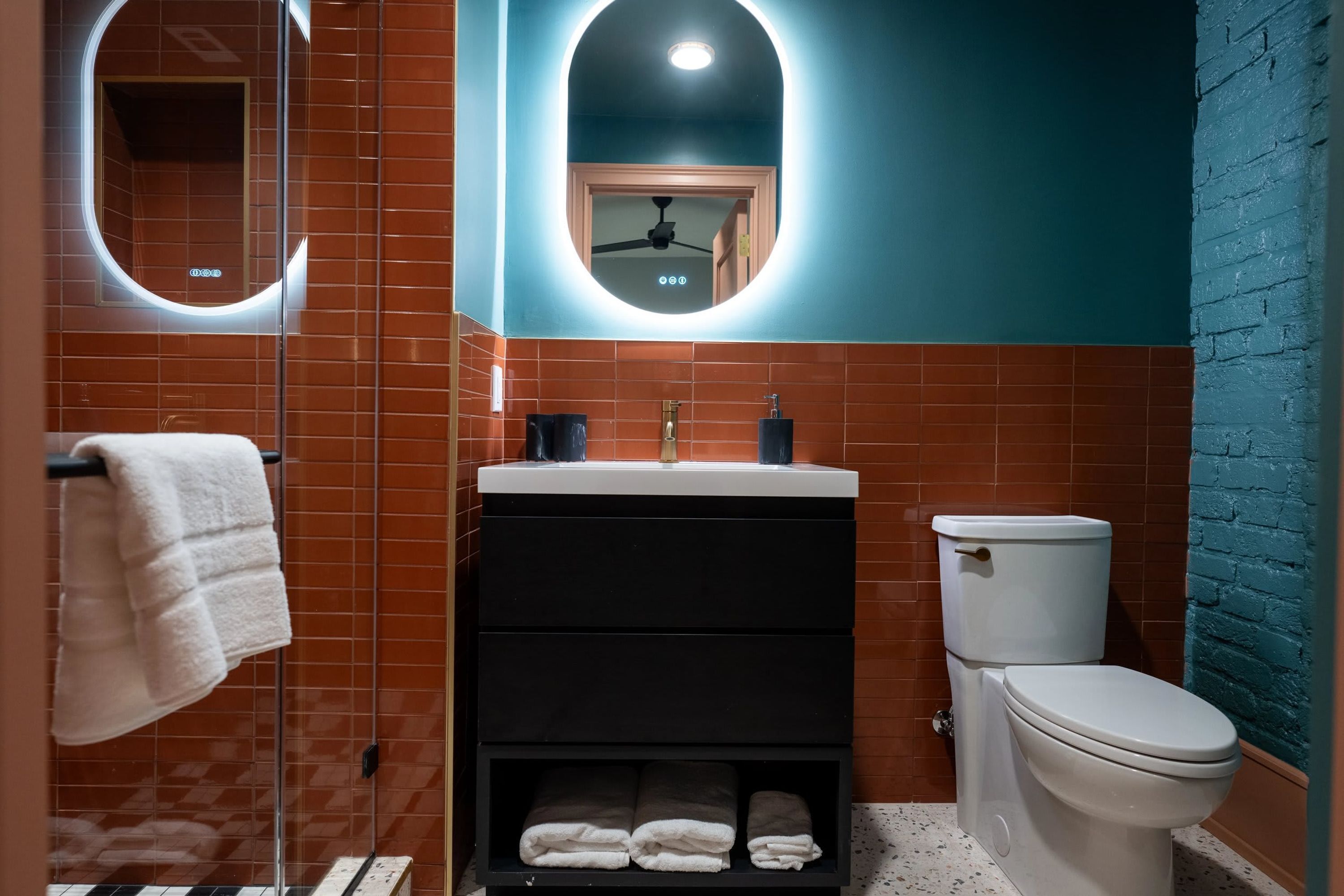 Une salle de bain avec des toilettes blanches, un lavabo noir et un miroir avec un éclairage intégré.