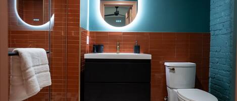 Une salle de bain avec des toilettes blanches, un lavabo noir et un miroir avec un éclairage intégré.