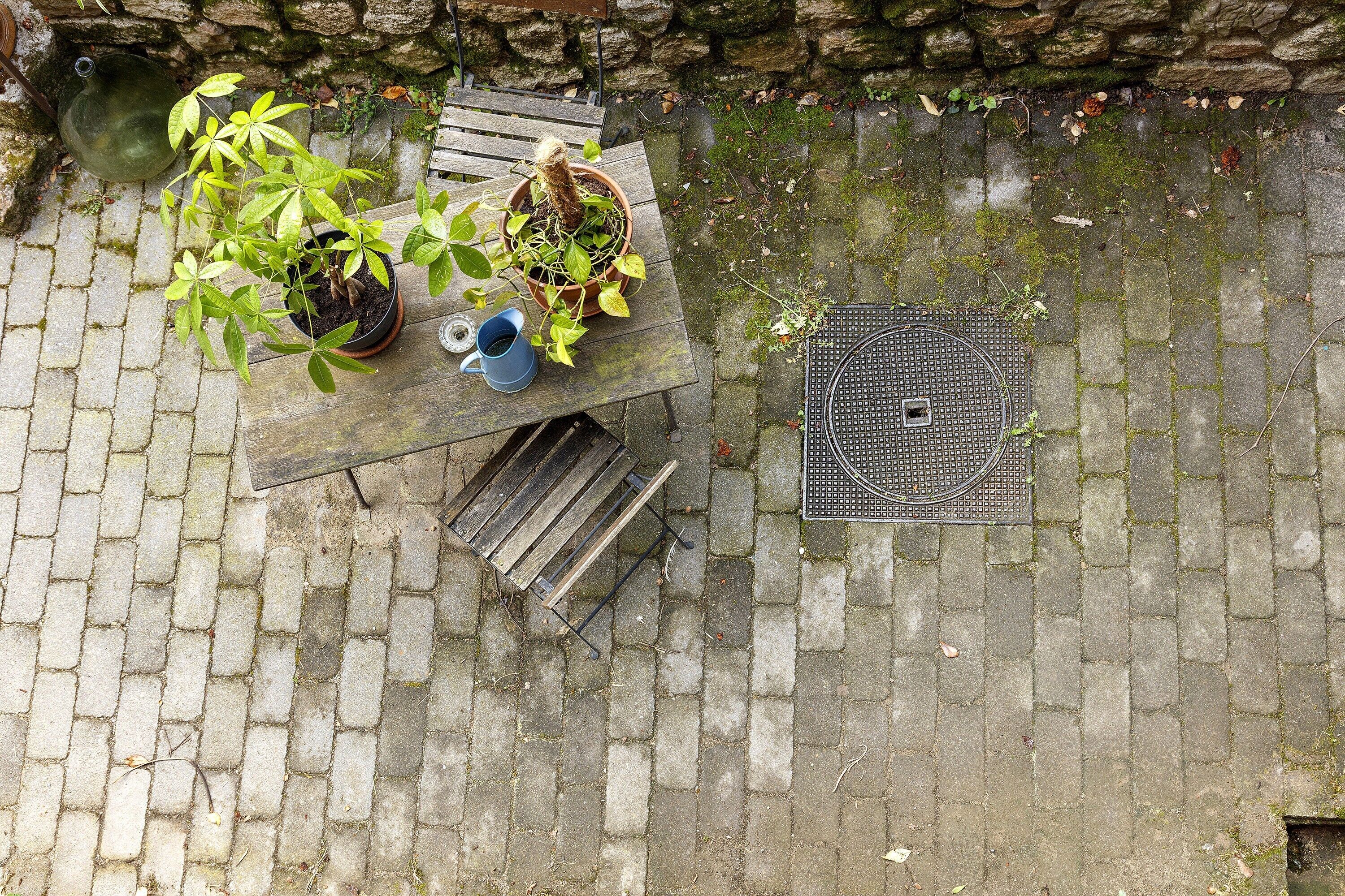 A patio with a wooden table, potted plants, a blue watering can, and a metal grate.