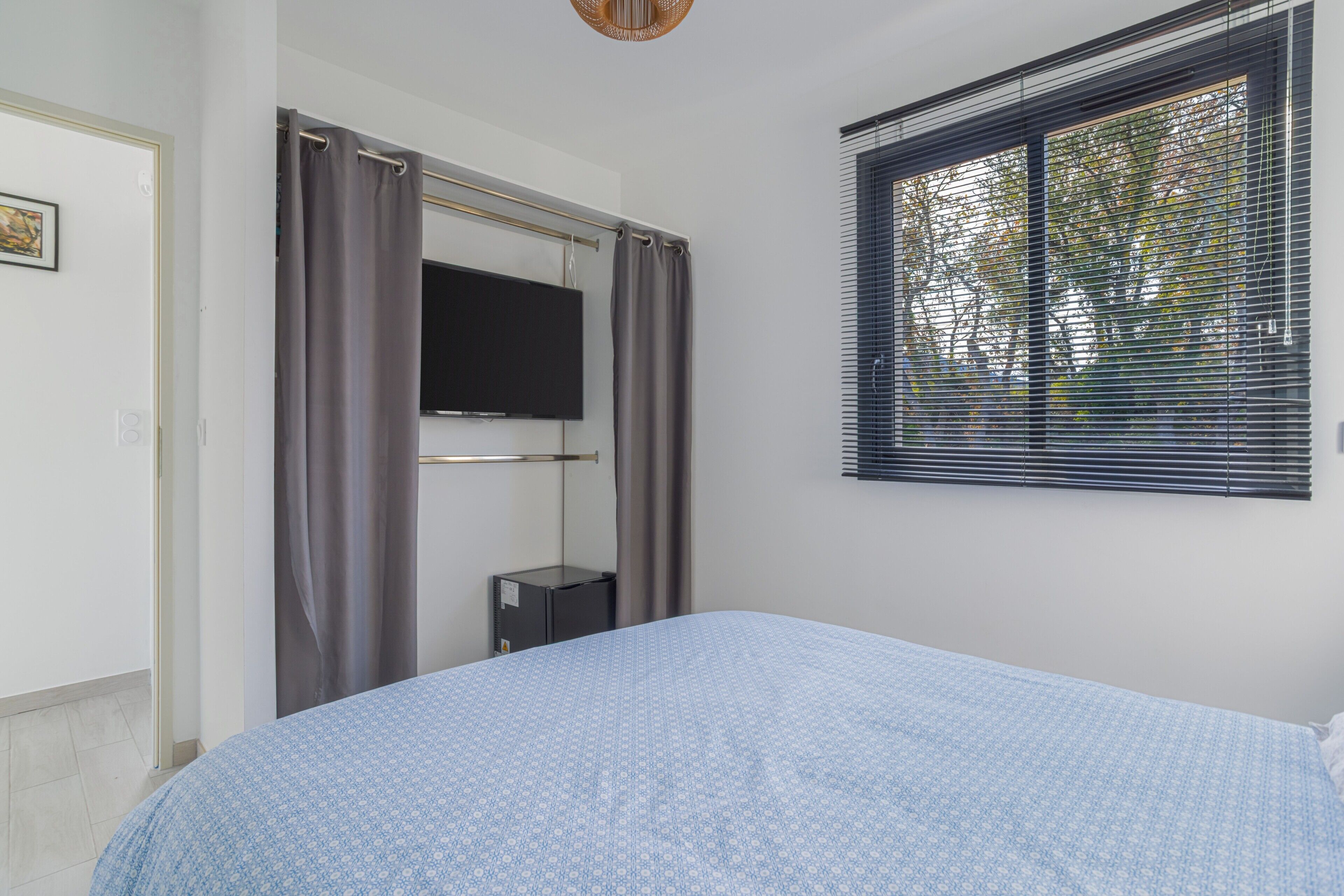 Une chambre à coucher comprenant un lit, un téléviseur, une fenêtre avec des stores et une tringle à rideaux.