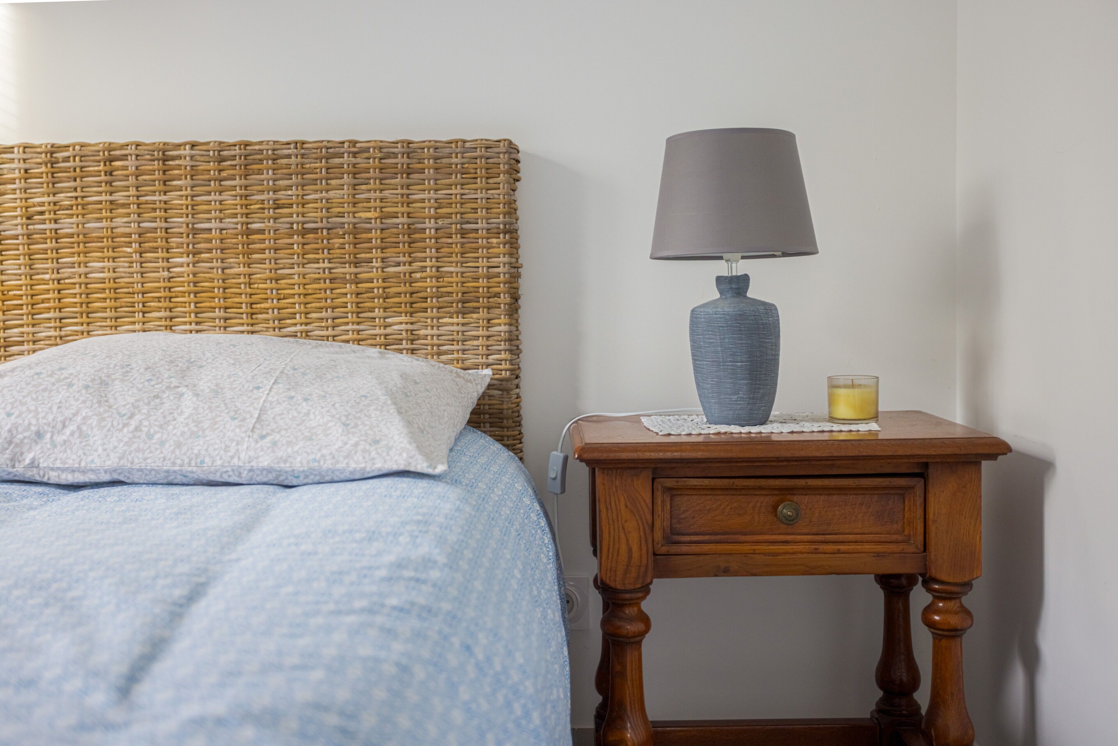 Een bed met een wit kussen en een blauwe deken, een houten nachtkastje met een lamp en een kaars, en een rieten hoofdbord.