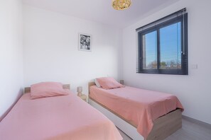 A bedroom with two single beds, a window with blinds, a wall-mounted picture, and a bedside table with a lamp.
