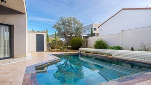 A modern swimming pool with a clear blue water, surrounded by a stone deck and a white wall.