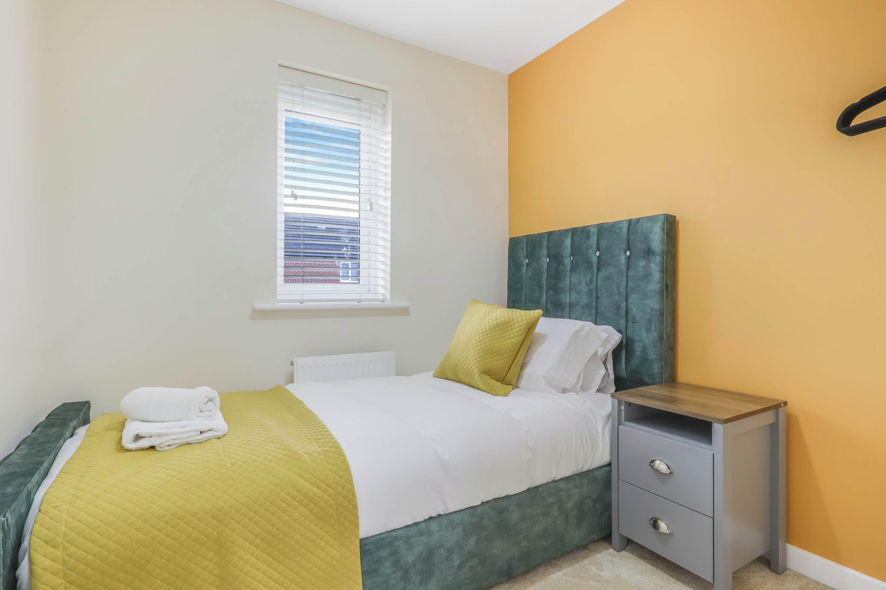 A neatly made bed with a green headboard, a bedside table with drawers, and a window with blinds.