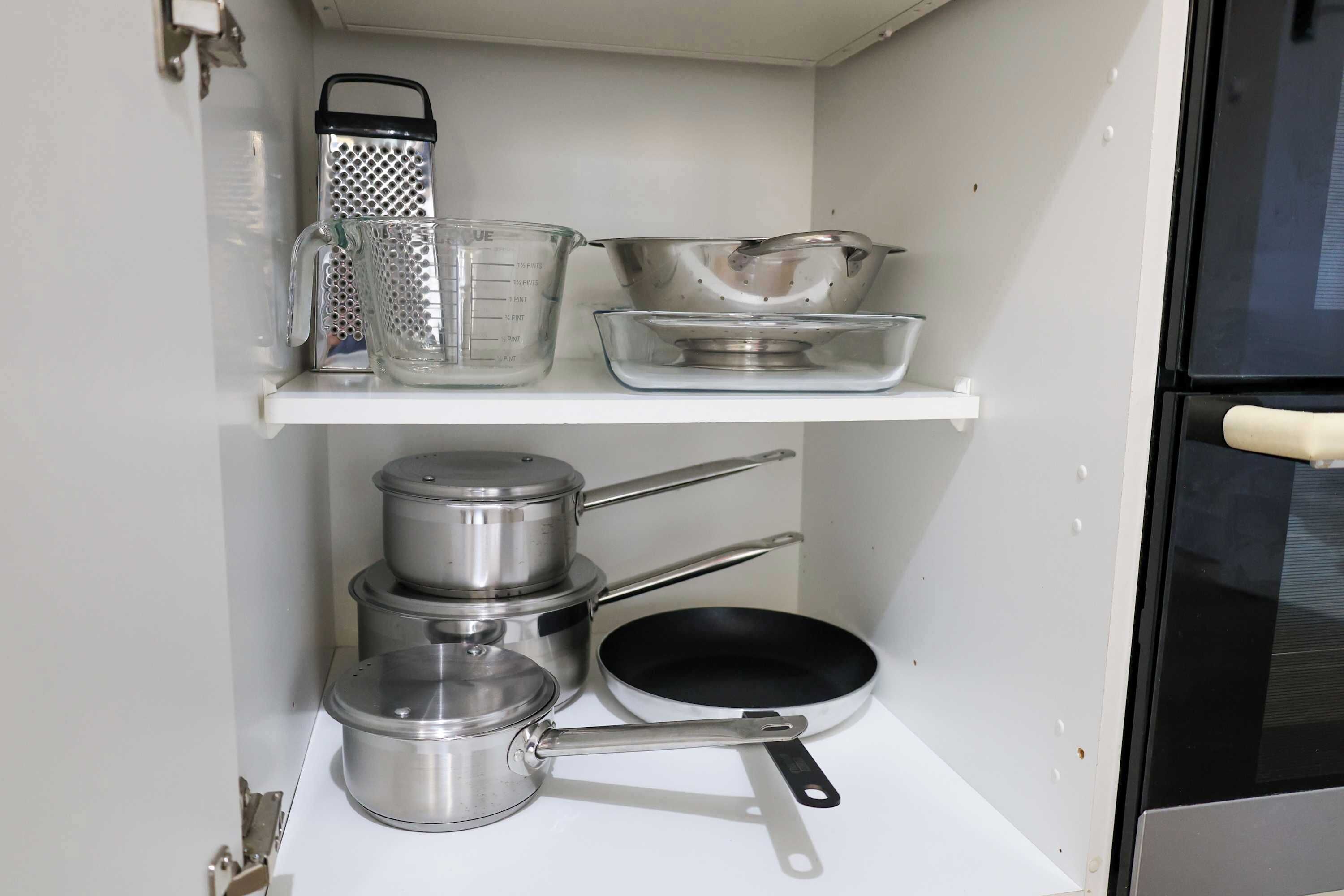 Ett kök med köksredskap i rostfritt stål, inklusive kastruller, stekpannor och ett rivjärn, placerade på en vit hylla.