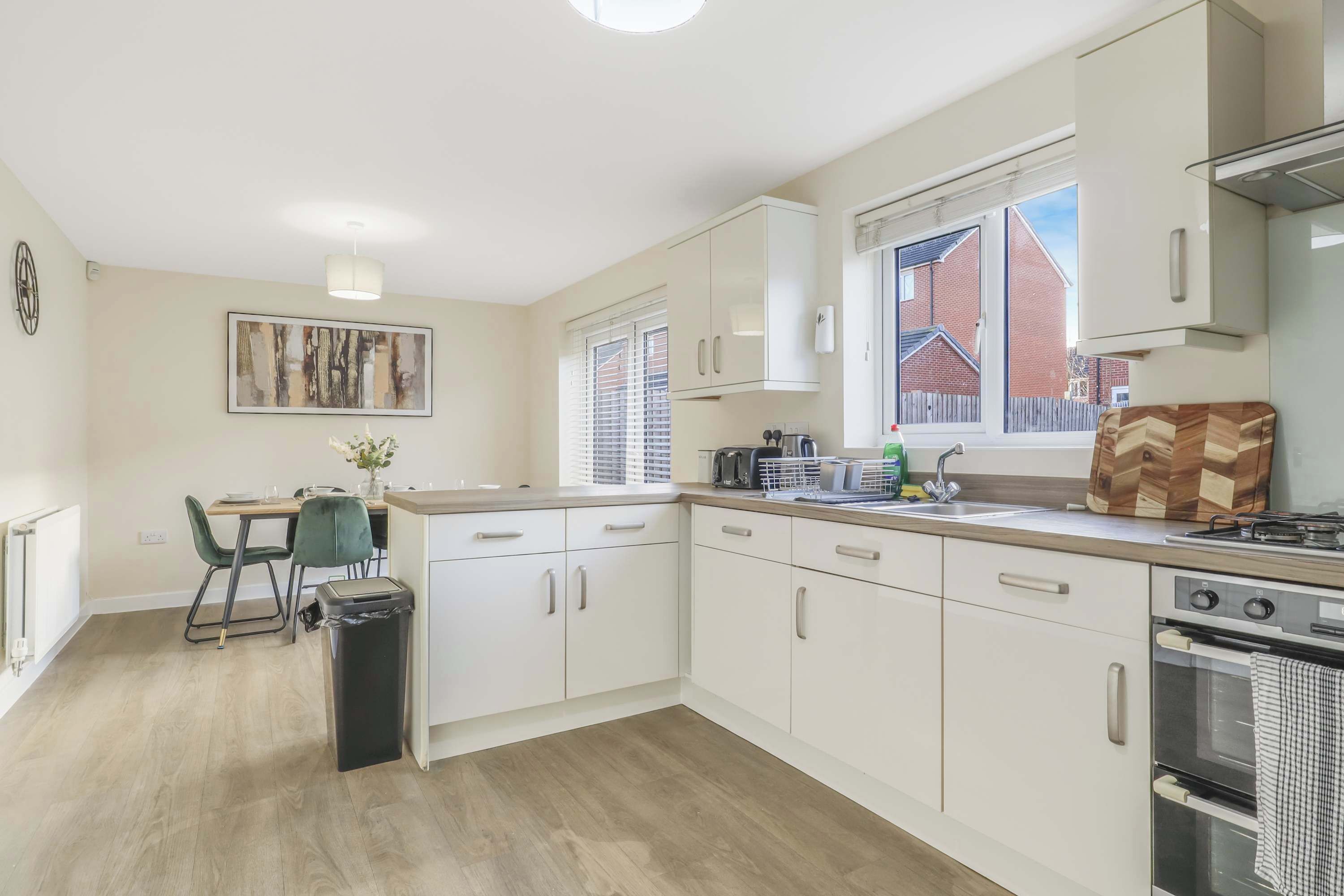 A modern kitchen with white cabinets, a dining area with a green chair, and a window with blinds.