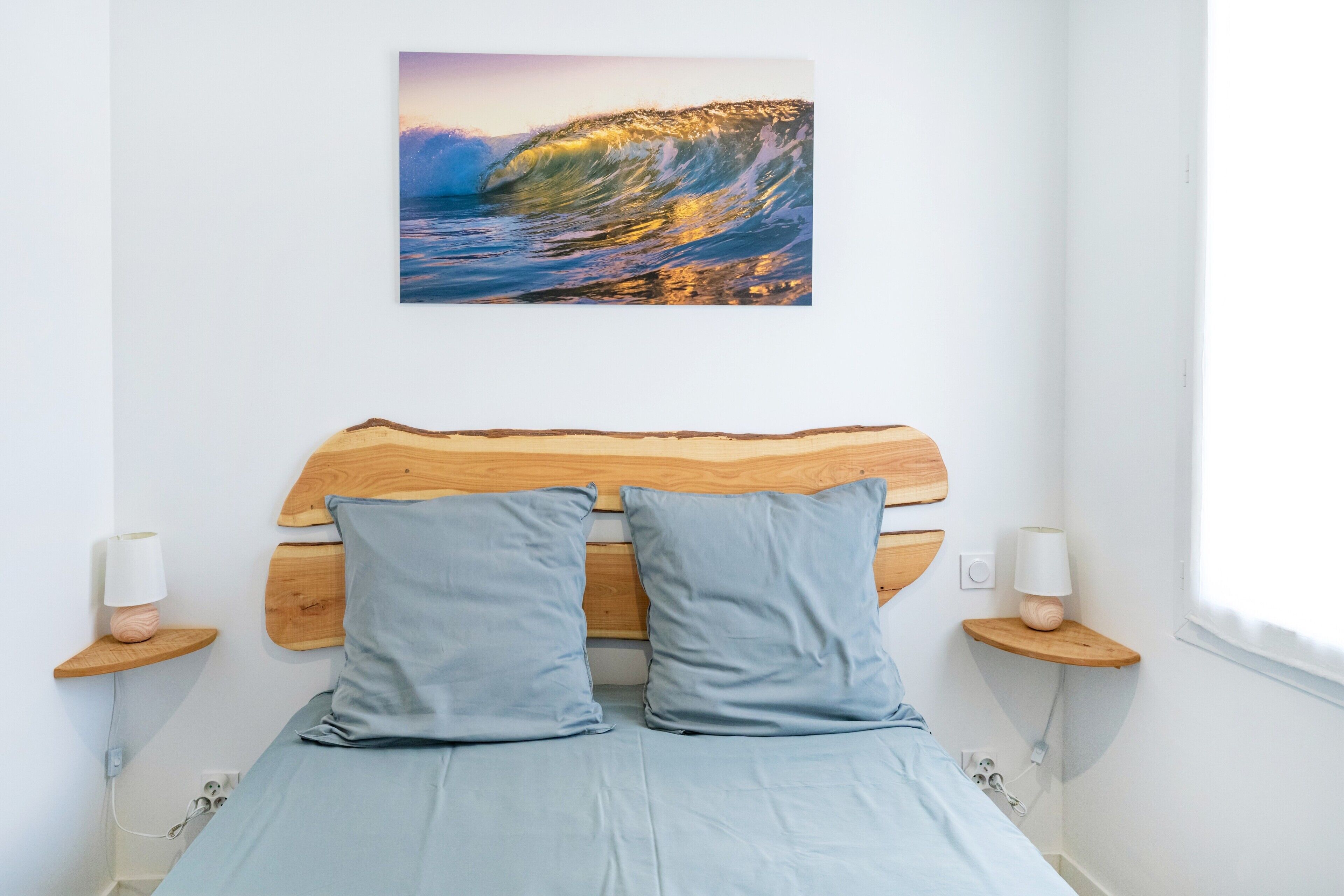 Un dormitorio con cabecera de madera, dos almohadas, lámparas de noche y un cuadro colgado en la pared que muestra una ola.
