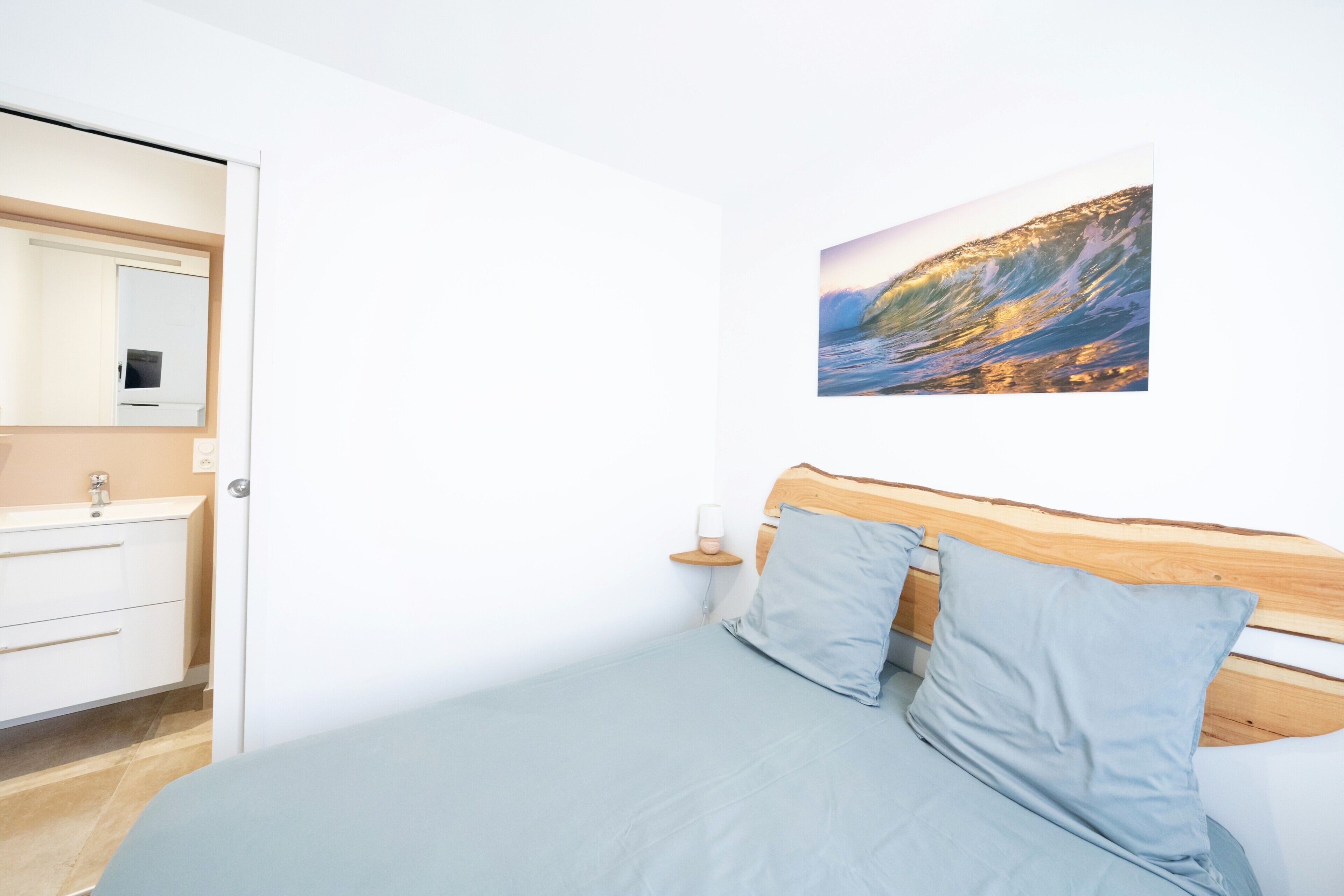 A bedroom with a bed, a wooden headboard, a nightstand with a lamp, a wall-mounted picture of a coastal scene, and a white wardrobe.