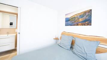 A bedroom with a bed, a wooden headboard, a nightstand with a lamp, a wall-mounted picture of a coastal scene, and a white wardrobe.