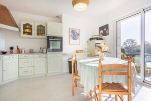 A kitchen with white cabinets, a dining table with a patterned tablecloth, and a view through a large window. - Windows to the Infinite Blue – Sea View & Direct Beach Access (Concarneau)