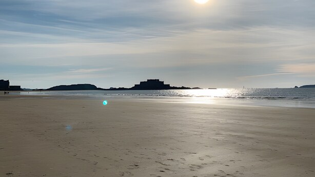 Une plage parsemée d’empreintes, un bâtiment lointain et le soleil qui se couche à l’horizon.