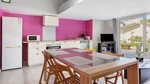 A modern kitchen with a pink wall, white cabinets, a glass dining table, and a TV in the corner.