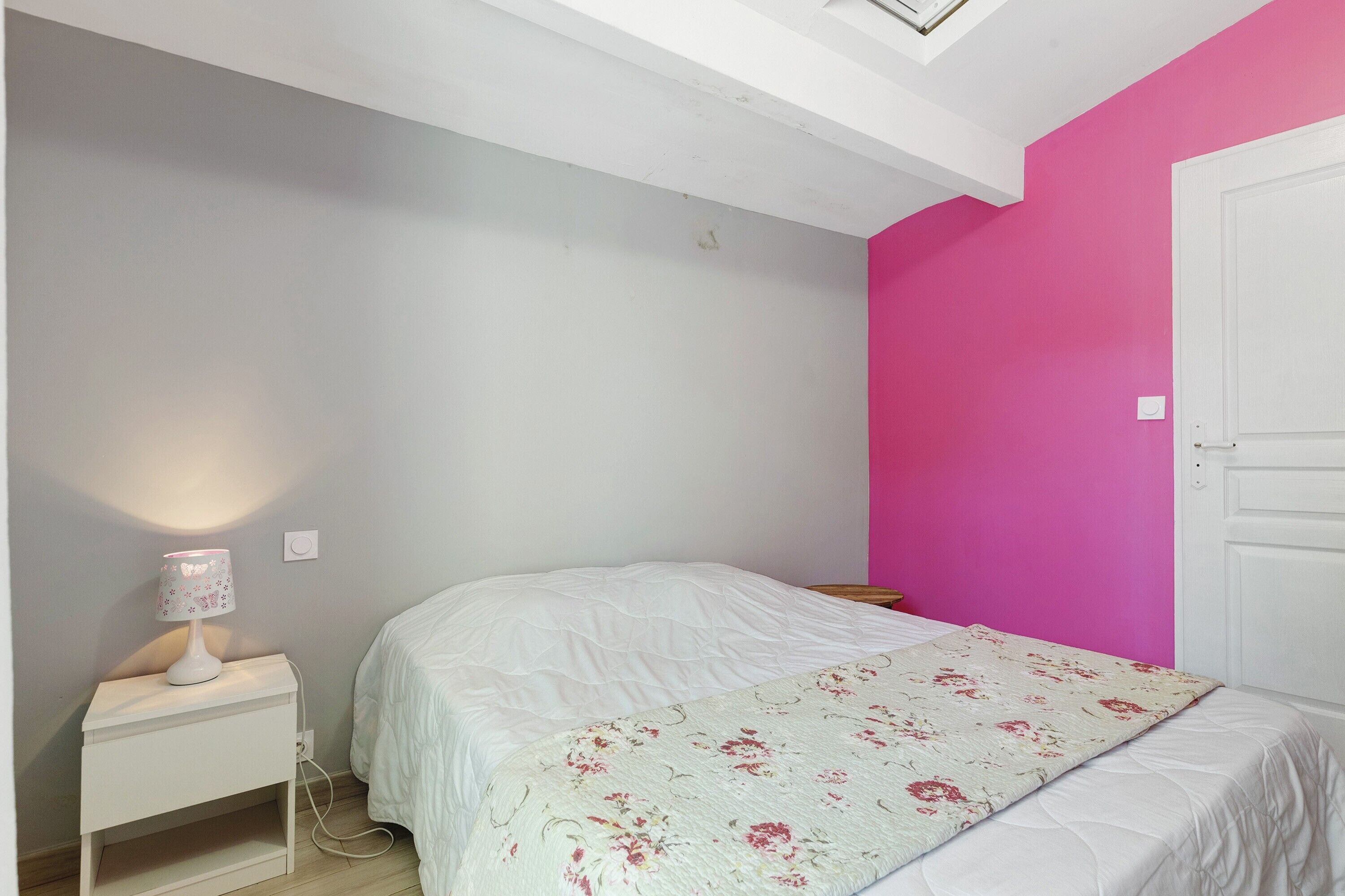 Ein Schlafzimmer mit einem Bett, einem Nachttisch mit Lampe, einer weißen Tür und einer rosafarbenen Wand.