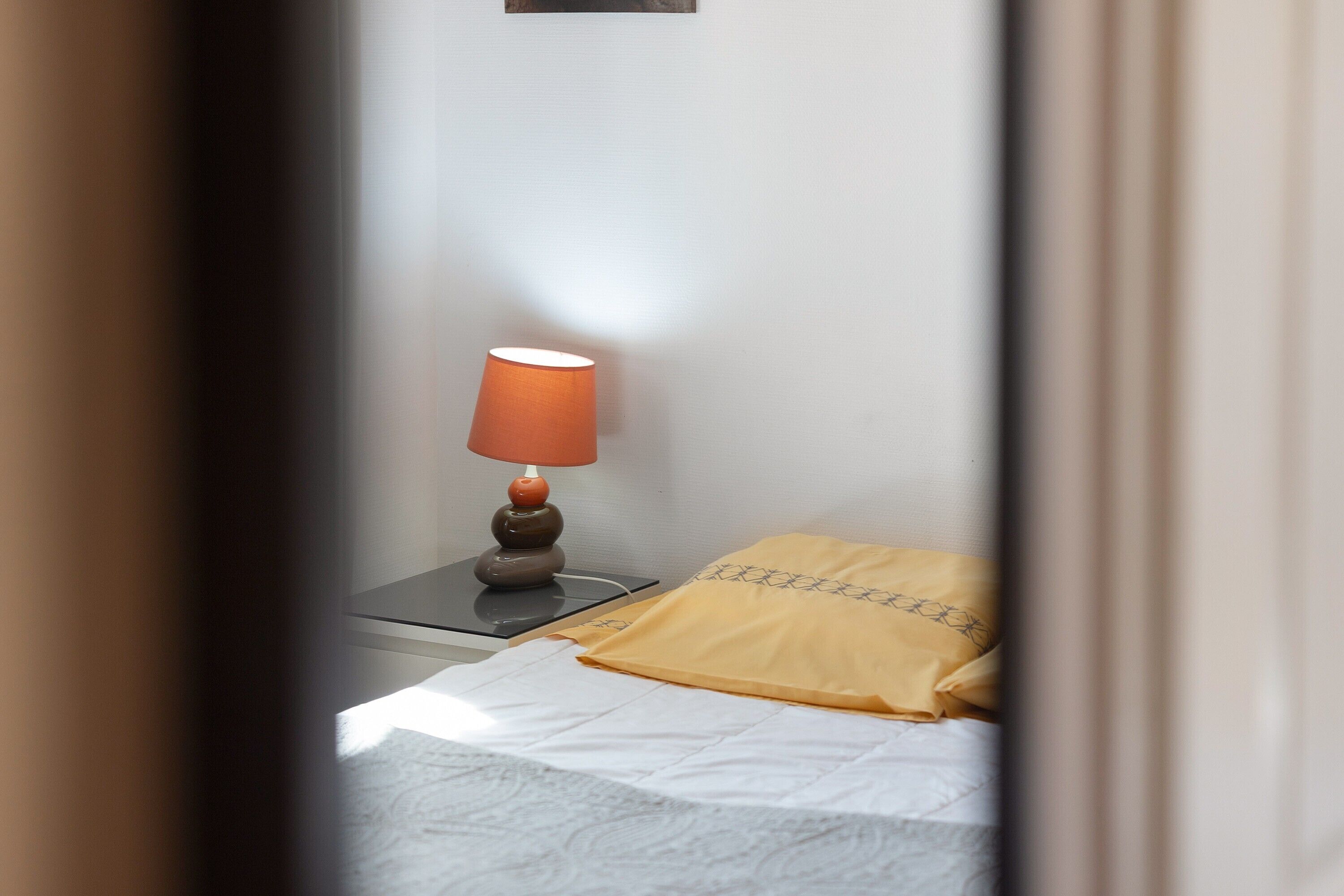 A bed with a white sheet and a yellow pillow, a bedside table with a lamp, and a wall-mounted light fixture.